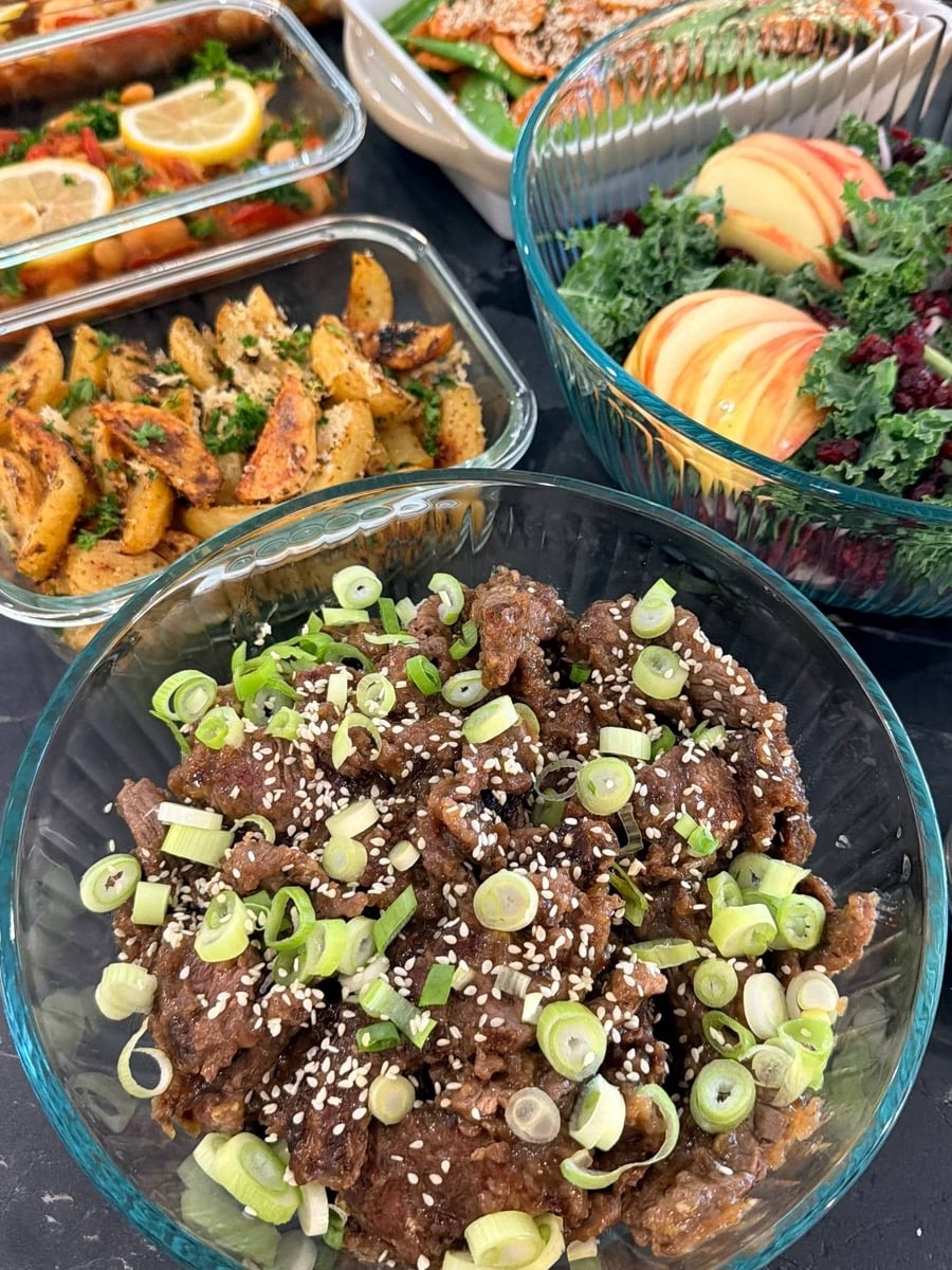 A glass bowl filled with tender teriyaki beef sirloin strips, garnished with fresh sliced scallions and toasted sesame seeds, served alongside varied meal sides.