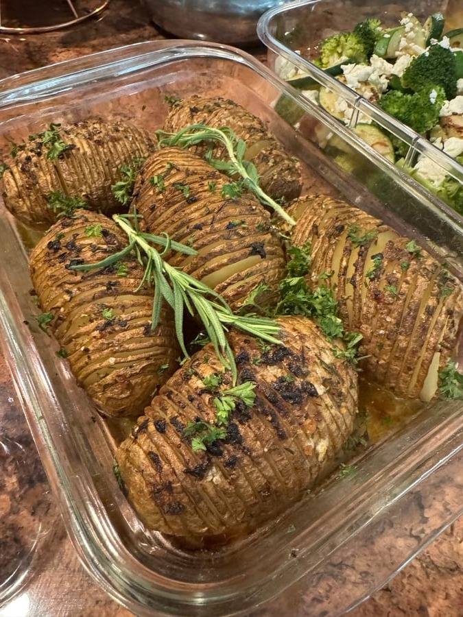 A glass container filled with golden, crispy hasselback potatoes seasoned with roasted garlic and fresh rosemary sprigs, alongside a side of sautéed broccoli and zucchini.
