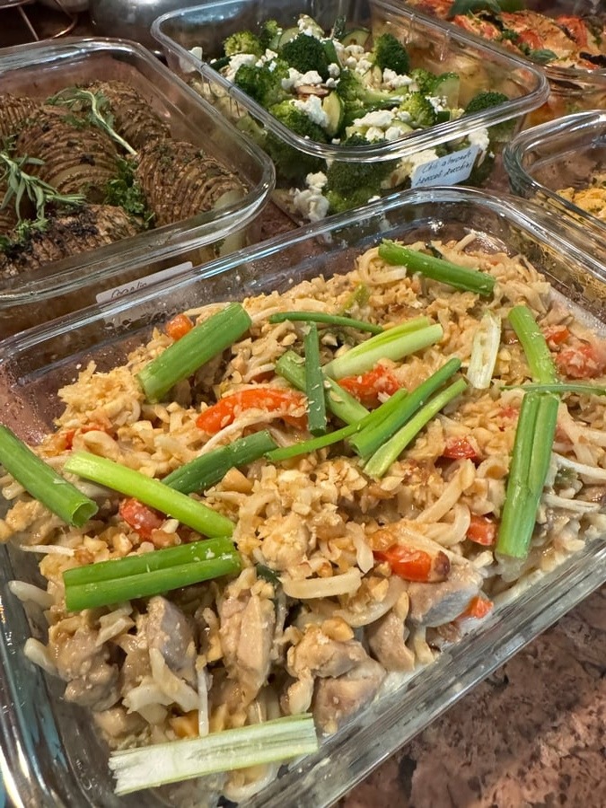 A glass container features chicken peanut Pad Thai noodles topped with crunchy crushed peanuts, vibrant sliced bell peppers, and fresh chopped green onions.