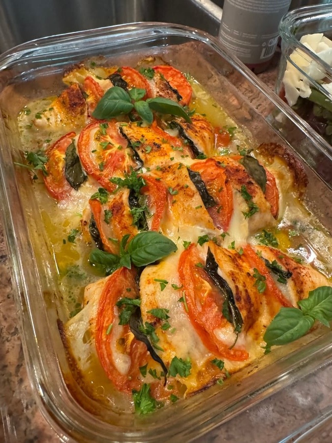 A glass baking dish containing tomato basil mozzarella chicken breasts, topped with fresh green basil leaves and herbs, served in a clear rectangular container.