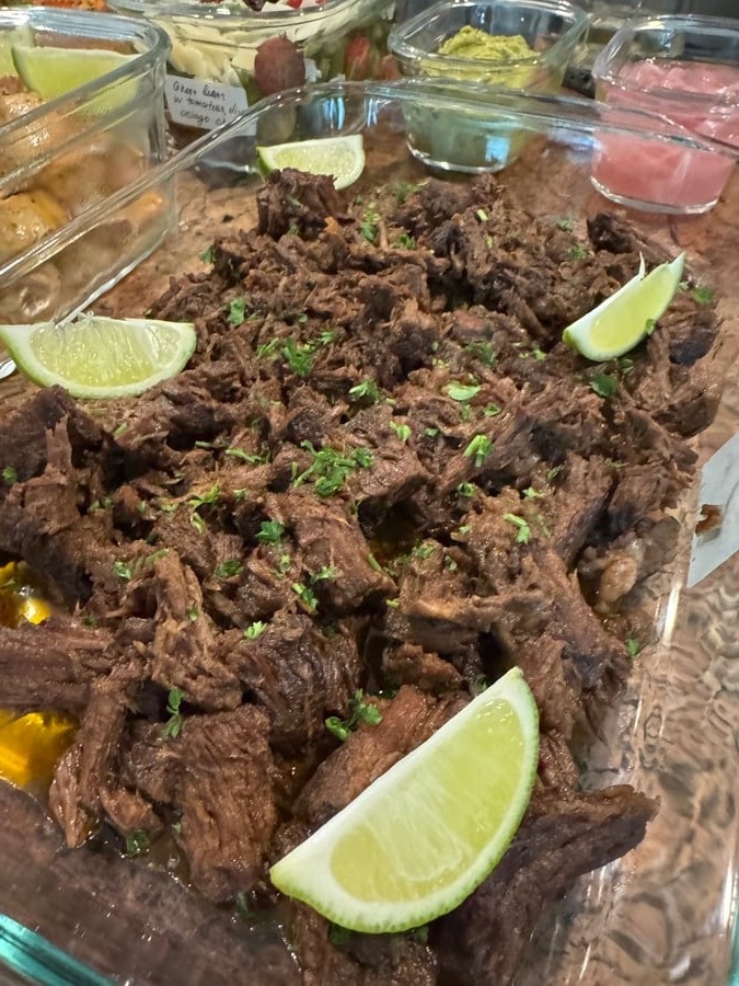 A large glass dish filled with tender Mexican style shredded beef garnished with fresh lime wedges and a sprinkling of chopped green herbs.