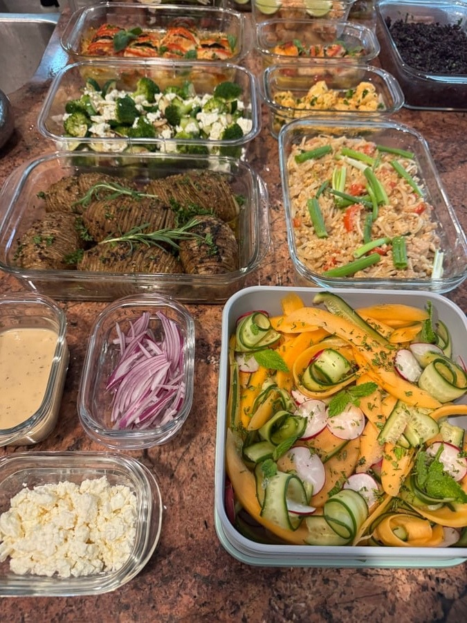 A variety of glass containers filled with hasselback potatoes, pad thai, broccoli zucchini, and melon cucumber salad, all neatly arranged for easy service.
