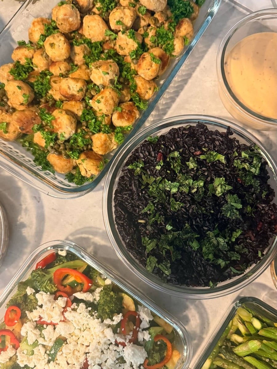 Glass containers featuring chicken picatta meatballs with capers, cilantro almond black rice, chili seared broccoli and zucchini, and ginger garlic asparagus stir-fry.