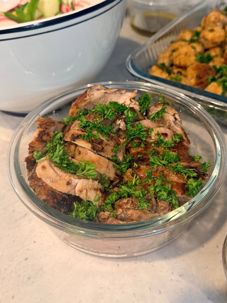 A glass bowl containing sliced pork tenderloin with maple mustard sauce, garnished with fresh parsley, alongside a bowl of melon salad and salmon bites.