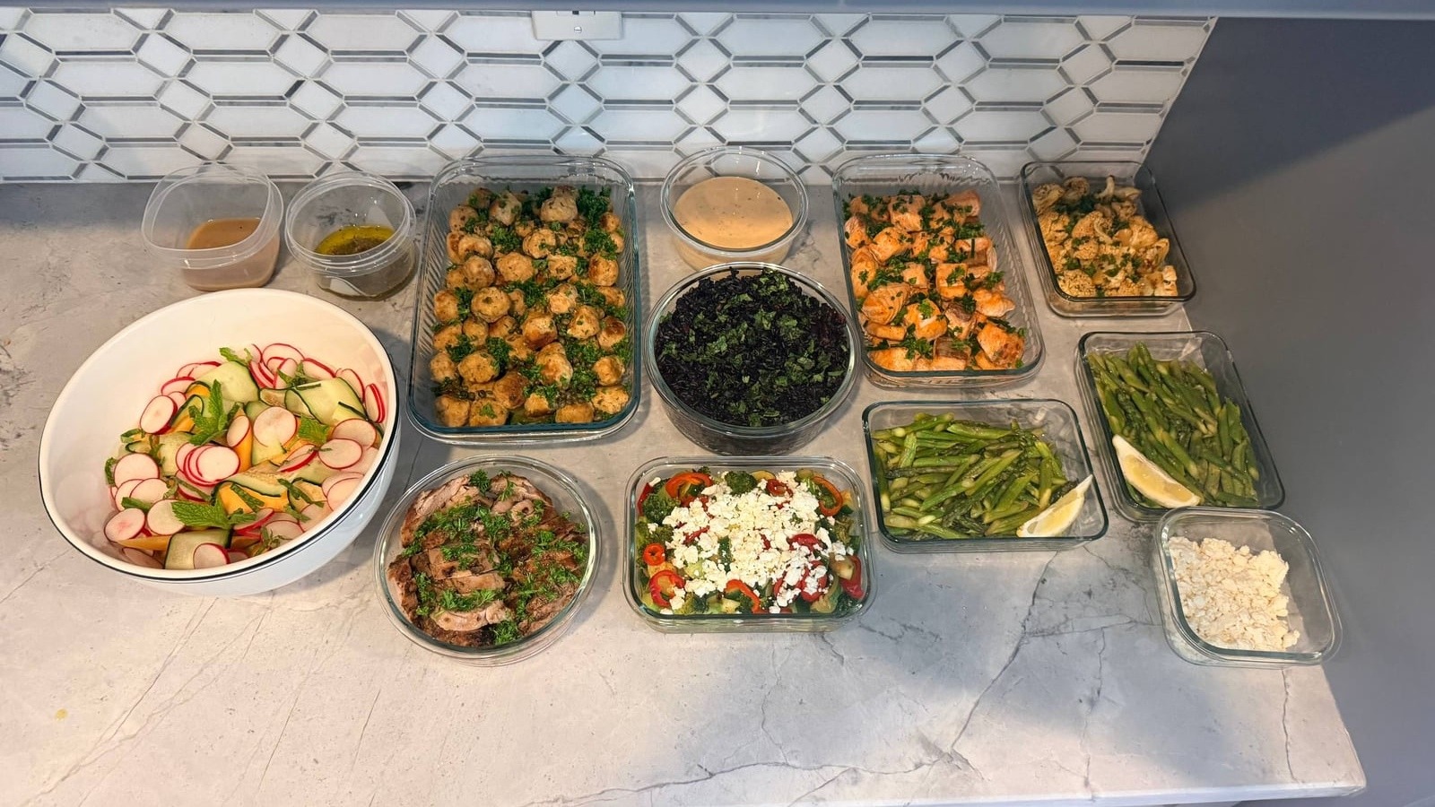 A variety of prepared dishes including chicken meatballs, garlic salmon, pork tenderloin, black rice, and a colorful salad served in glass containers.