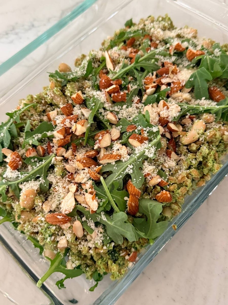 A glass dish filled with vibrant green arugula pesto farro and cannellini beans, topped with fresh arugula leaves, toasted almonds, and a dusting of parmesan cheese.