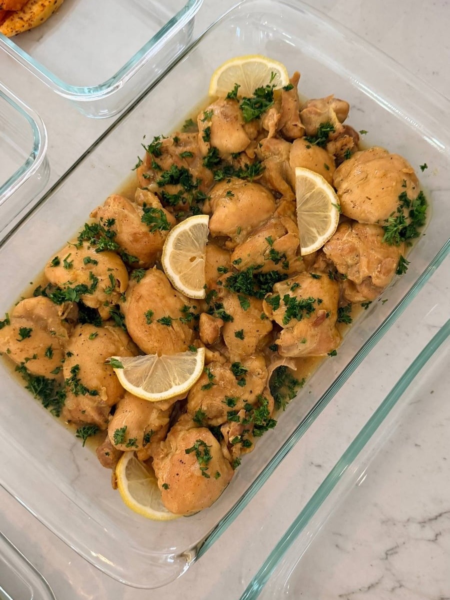 A large glass dish filled with golden saffron turmeric baked chicken thighs garnished with fresh lemon slices and finely chopped green parsley.