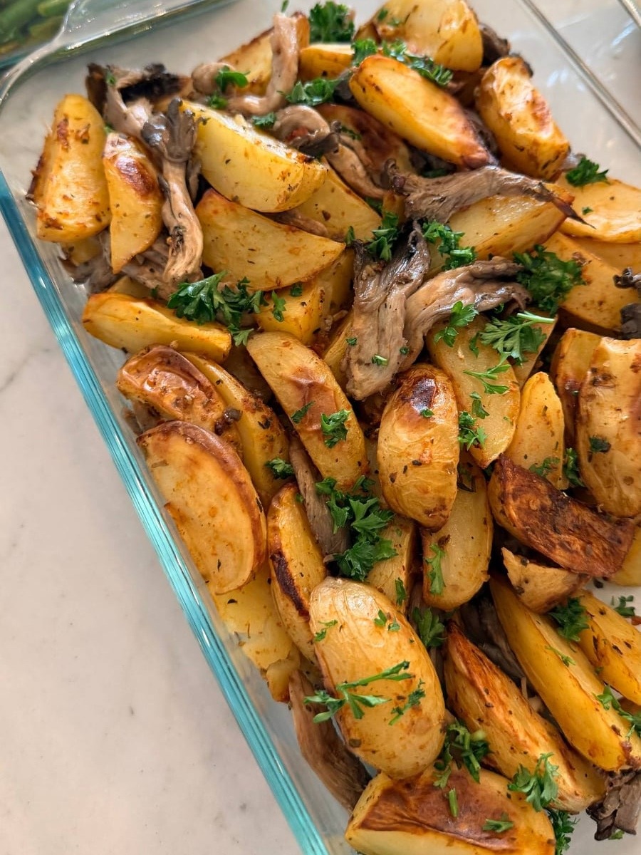 A glass container features golden-brown baked fingerling potatoes and savory oyster mushrooms, garnished with fresh green parsley on top of the roasted vegetables.