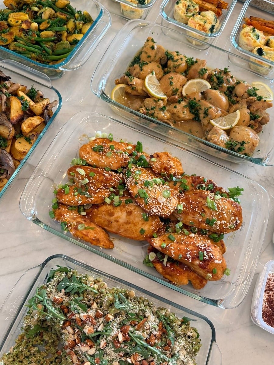 An overhead view of multiple glass containers filled with honey garlic chicken, saffron chicken, arugula pesto farro, and assorted vegetable sides on a counter.
