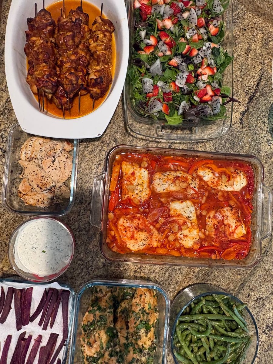 A variety of meal prep containers featuring bbq chicken skewers, harissa cod with butter beans, a strawberry dragon fruit salad, and lemon green beans.