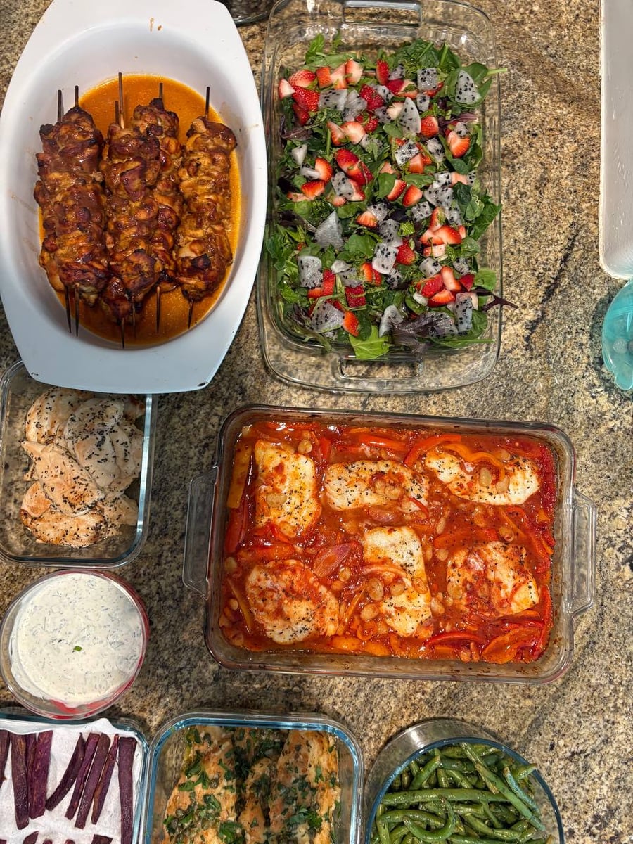 Top-down view of organized food containers featuring BBQ chicken skewers, a vibrant fruit salad, harissa-baked cod, and sides of green beans and purple potatoes.