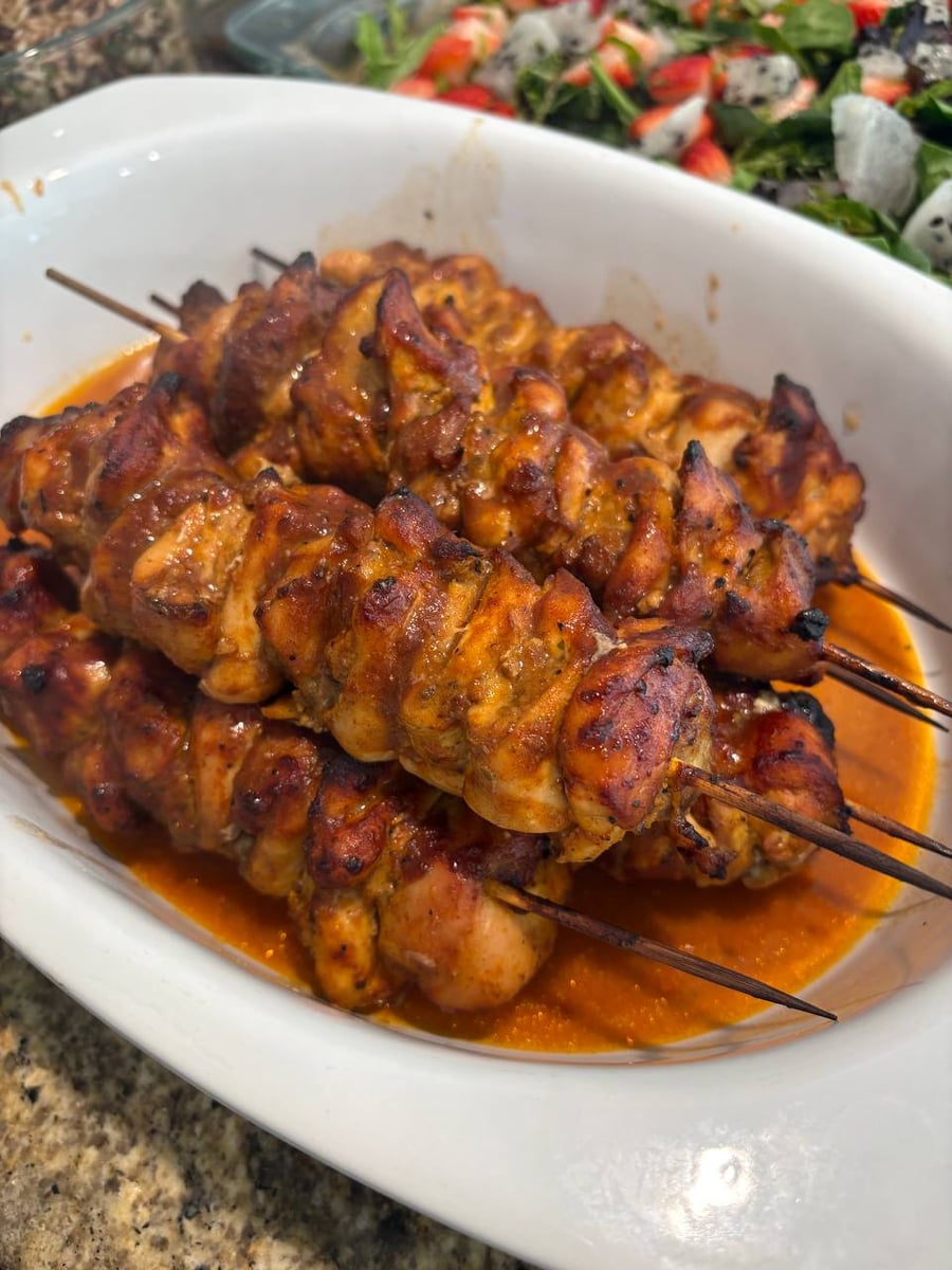 A white ceramic casserole dish holding BBQ-glazed chicken thigh skewers, served alongside a salad with dragon fruit, strawberries, and greens in the background.