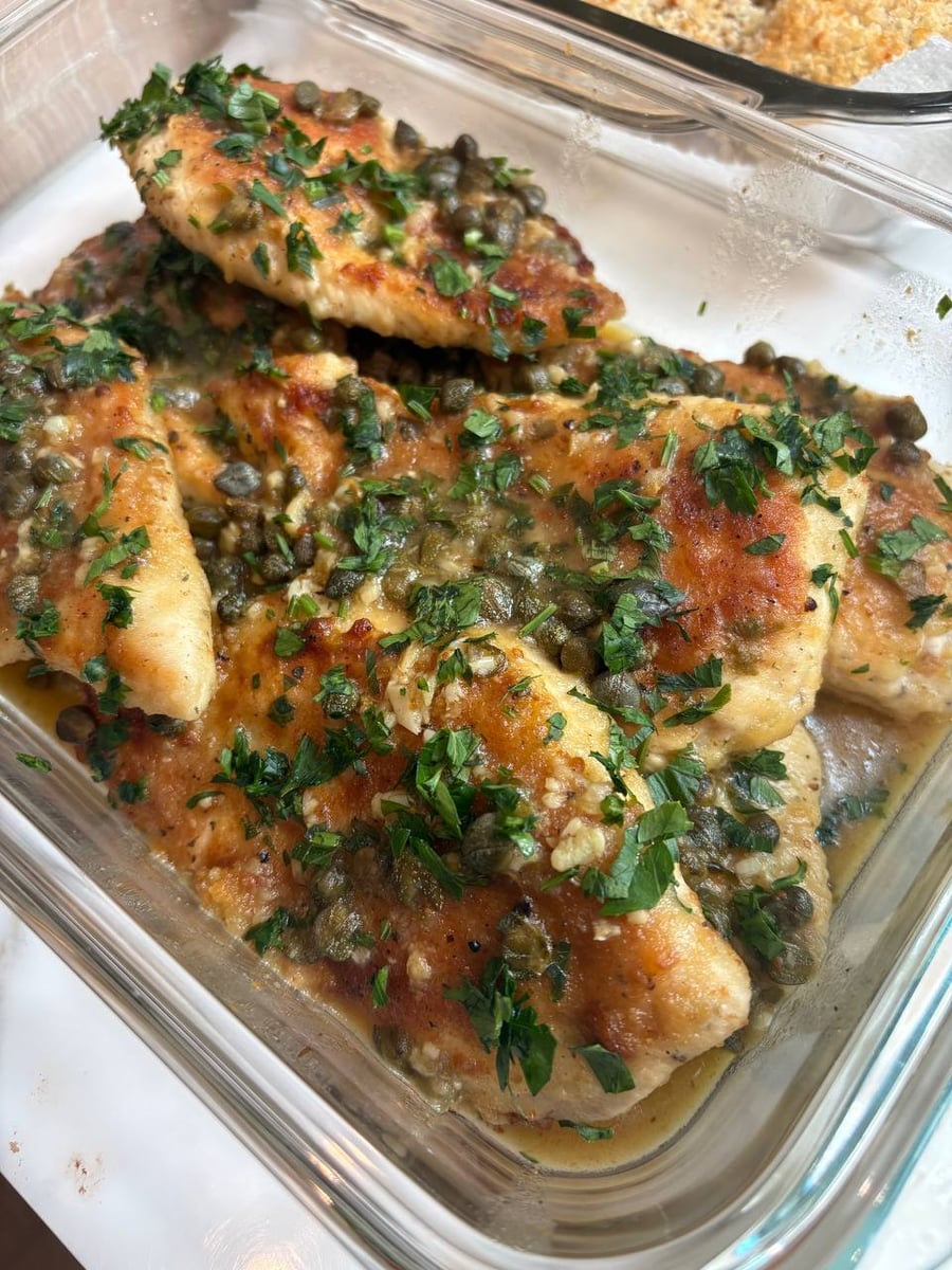 A glass container filled with seared chicken breast piccata, generously topped with tangy capers, fresh parsley garnish, and a zesty lemon butter sauce.