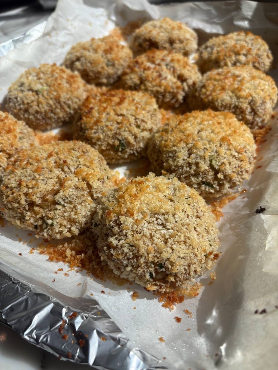A baking sheet lined with parchment paper holds several golden-brown crispy baked crab cakes coated in coarse, toasted breadcrumbs arranged in a neat grid.
