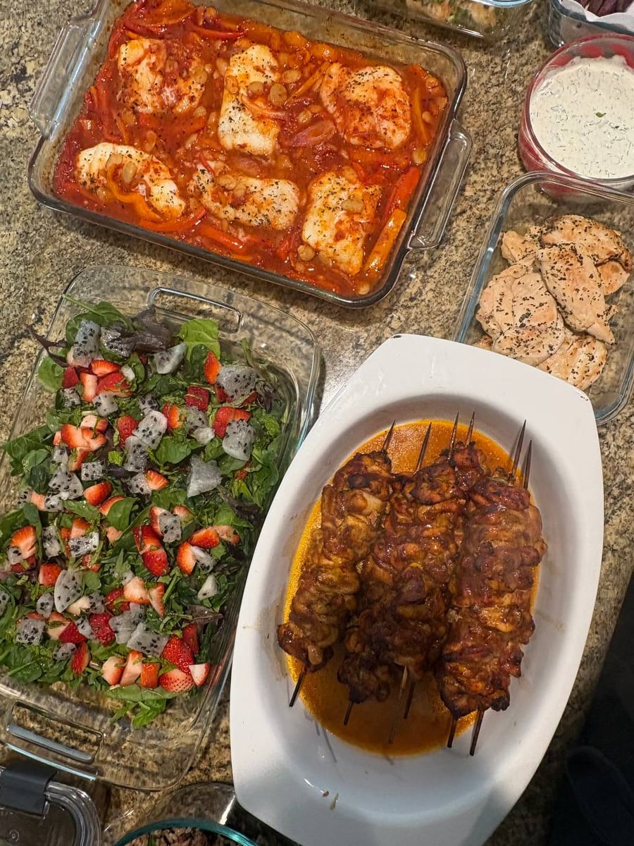 A top-down view of food preparations including harissa baked cod with beans, BBQ glazed chicken skewers, and a vibrant dragon fruit strawberry salad.