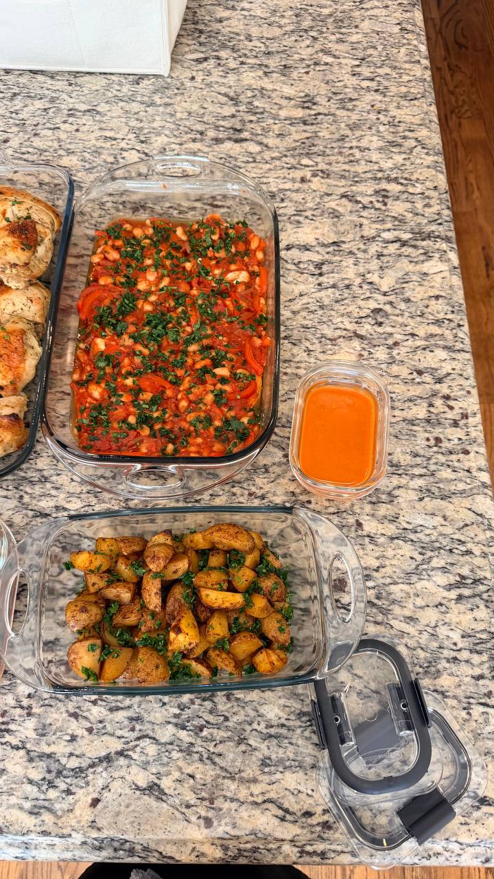 A glass baking dish filled with harissa cod, chickpeas, and bell peppers next to a dish of golden garlic herb roasted potato wedges with a side sauce.