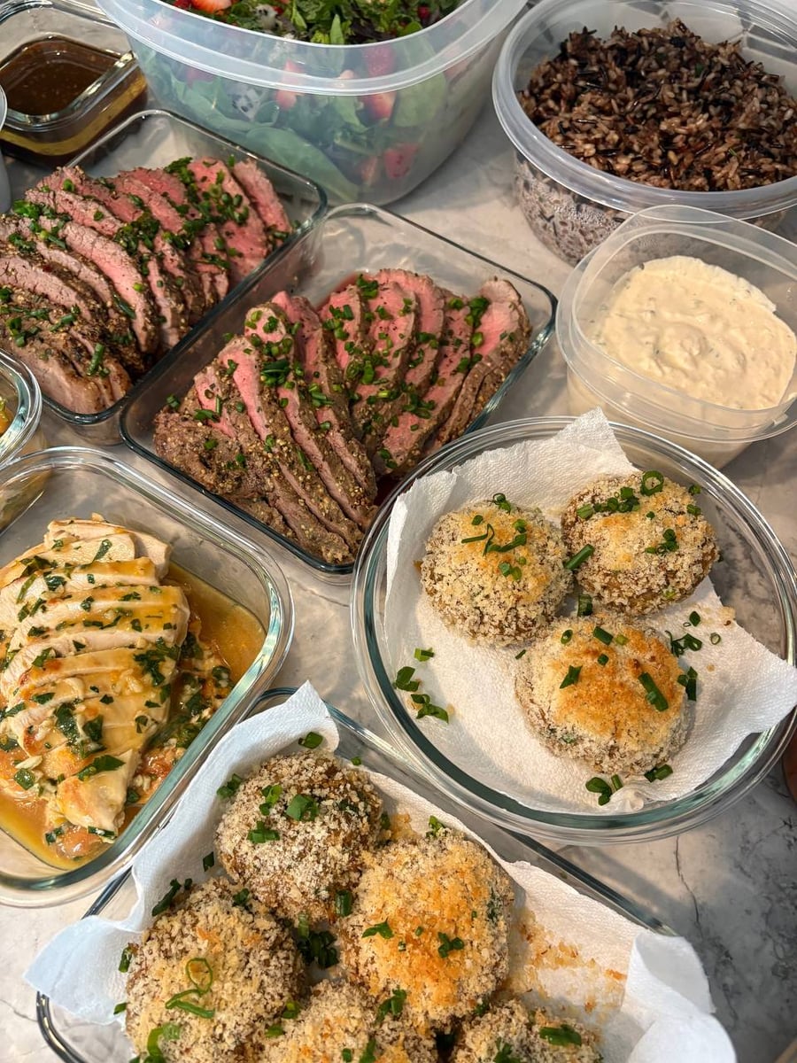 Glass containers filled with rosemary beef roast, orange glazed chicken breast, and crispy crab cakes with remoulade, side dishes, and a fresh salad.