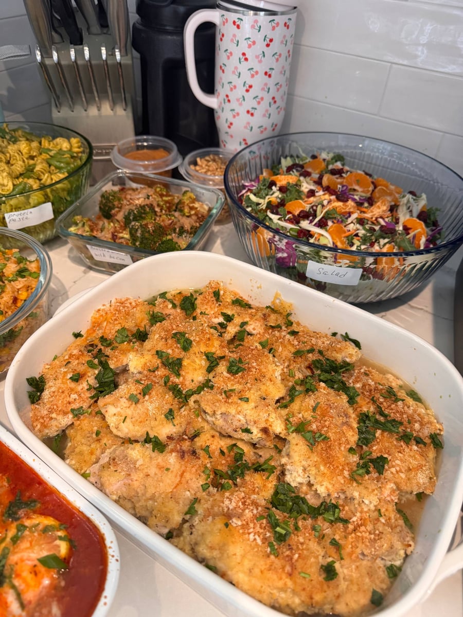 A large white casserole dish filled with crispy baked cheddar chicken thighs, topped with fresh parsley, served alongside colorful meal prep sides in a kitchen.