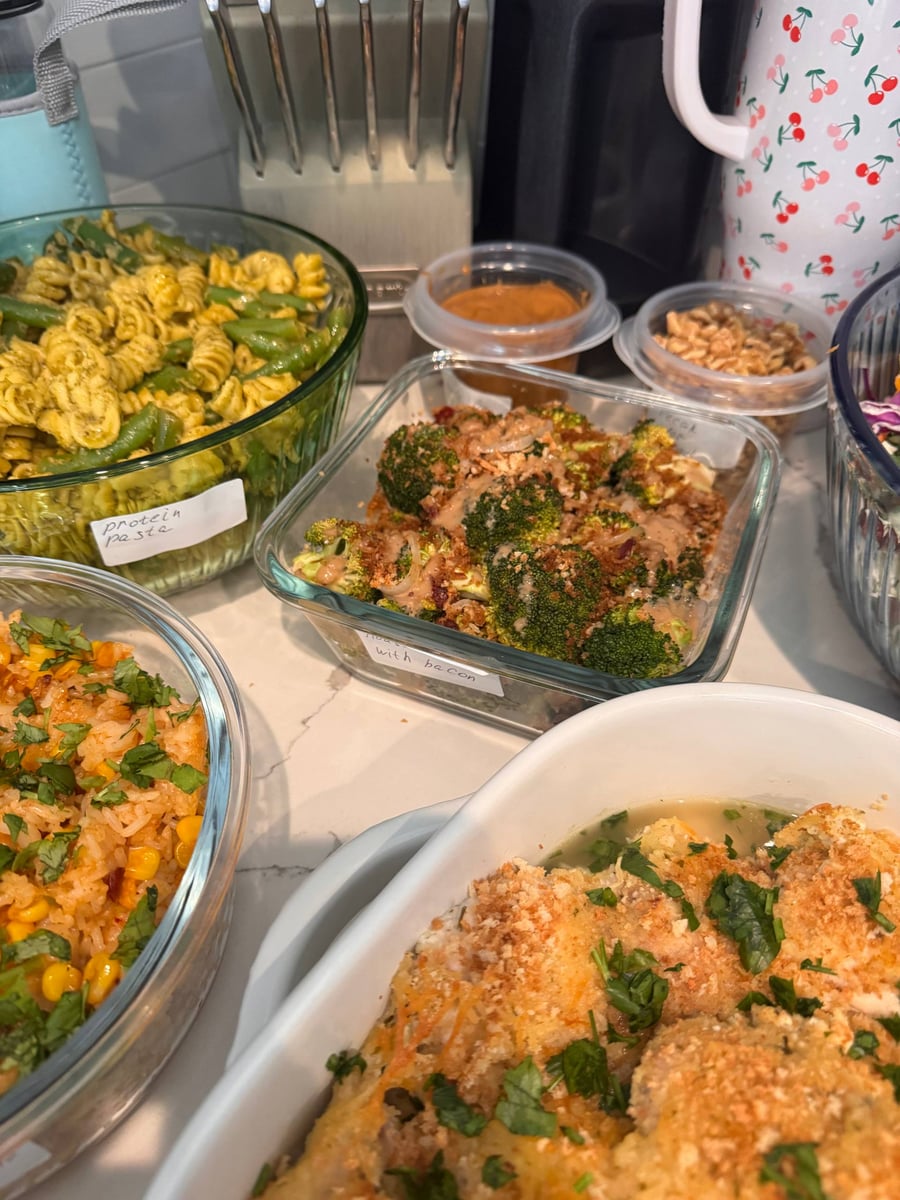 Glass containers holding crispy cheddar chicken, roasted broccoli with bacon, and pesto fusilli pasta, organized on a countertop for easy meal service.