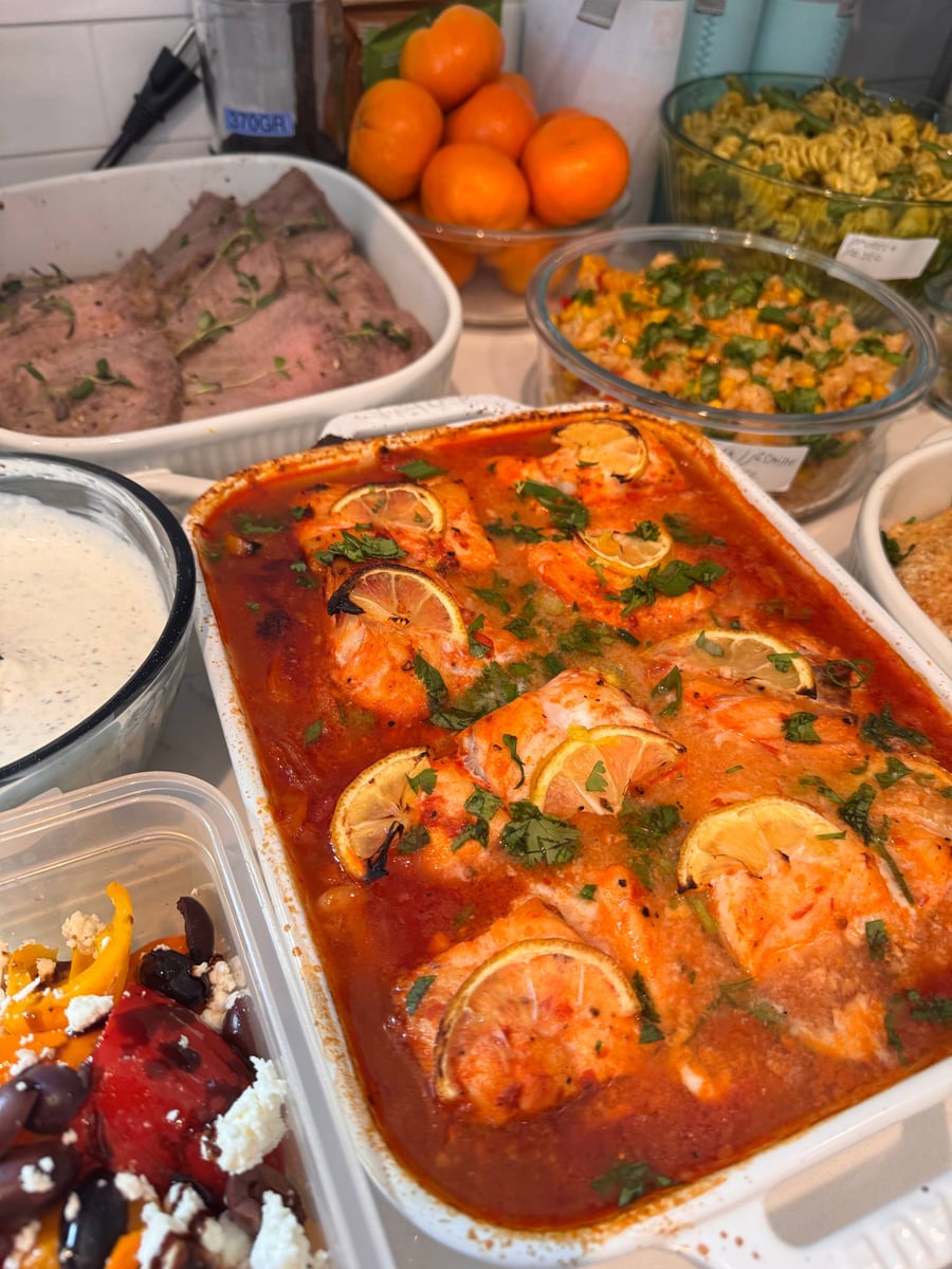 A baking dish filled with harissa butter beans and baked cod fish fillets, garnished with fresh lemon slices and fresh herbs.