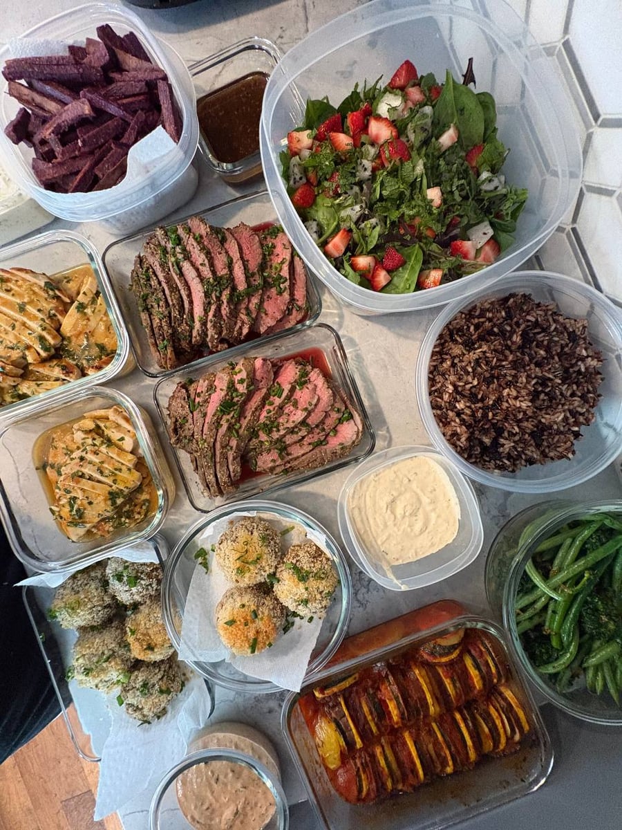 A collection of glass and plastic containers featuring sliced beef roast, orange chicken, crab cakes, ratatouille, purple sweet potato fries, and a fresh salad.
