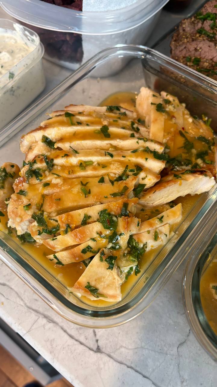 A glass container holds thinly sliced orange glazed chicken breast, topped with fresh chopped herbs and a shimmering citrus sauce for a vibrant presentation.