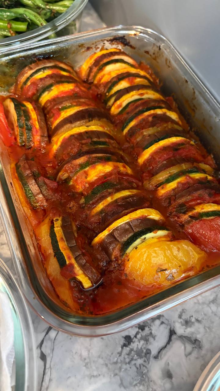 A glass baking dish contains beautifully arranged, thinly sliced ratatouille featuring layered zucchini, yellow squash, and eggplant nestled in a rich tomato base.