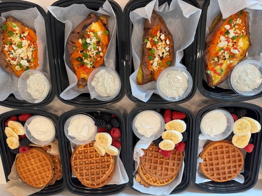 Nutritious breakfast meal prep containers featuring scrambled egg stuffed sweet potatoes with yogurt sauce and protein waffles with fresh berries.