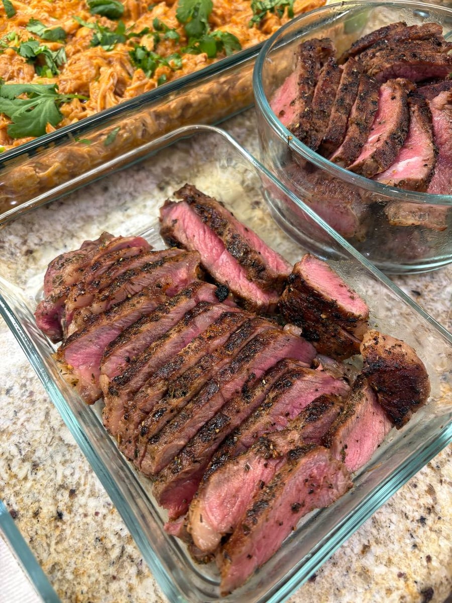 A glass container of herb-crusted, medium-rare sliced NY strip steaks sits beside a second glass container of saucy, cilantro-topped shredded chicken.