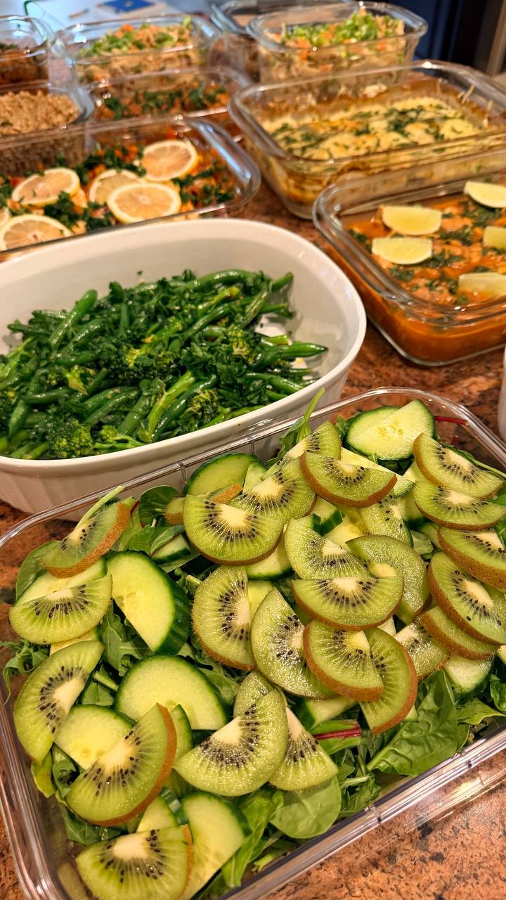 A glass container features a fresh salad of kiwi slices, cucumber, and greens, alongside a white bowl of vibrant chili crisp French beans and broccolini.