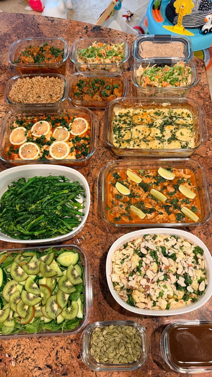 A variety of meal prep containers featuring saucy chicken, Tuscan meatballs, potato zucchini gratin, white fish, leafy salads, and roasted vegetables set on stone.
