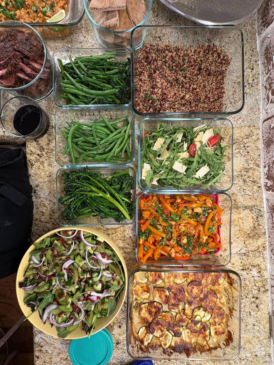 An assortment of glass meal prep containers featuring steak, herb-crusted chicken, buttery beans, roasted gratin, and a fresh kiwi-cucumber-spinach pumpkin seed salad.