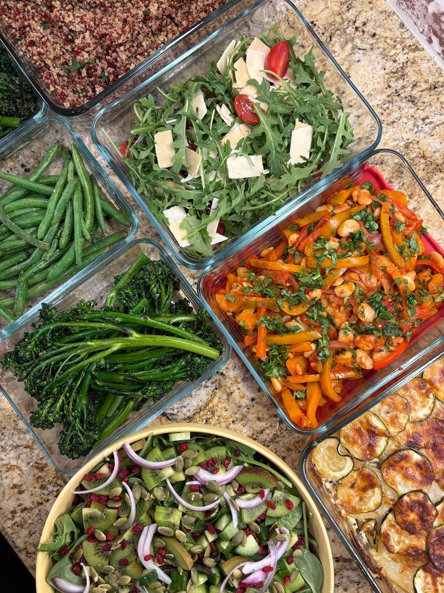 An overhead view of glass meal prep containers filled with butter bean stew, French gratin, fresh arugula salad, herb-crusted green beans, and kiwi salad.