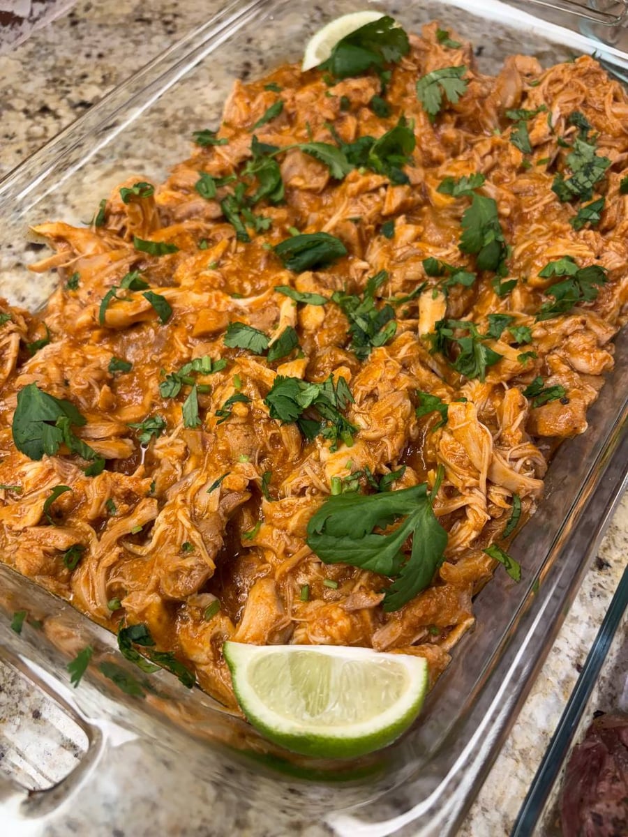 A glass baking dish filled with saucy shredded chicken, garnished with fresh cilantro leaves and two vibrant green lime wedges for a bright finish.