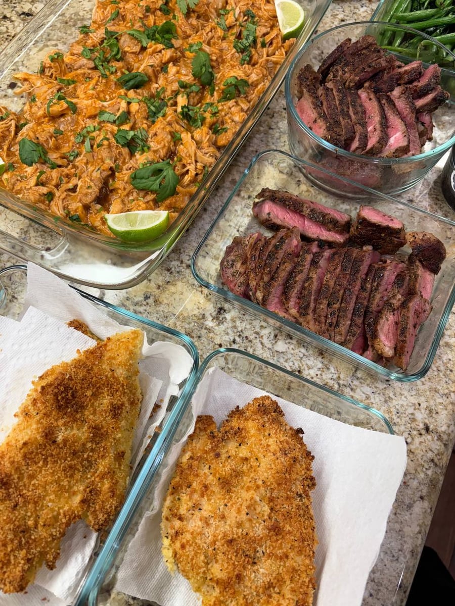 A spread of meal-prepped dishes including saucy shredded chicken, sliced medium-rare herb-crusted New York strip steaks, and crispy breaded Chicken Milano cutlets in glass containers.