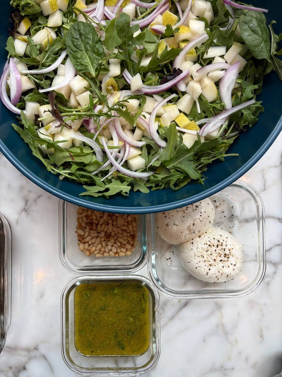 Peppery arugula salad base styled with creamy burrata cheese, toasted pine nuts, and red onion, representing high-end private chef meal prep aesthetics.
