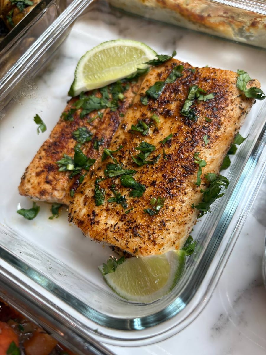 A glass meal prep container containing fillets of blackened mahi mahi seasoned with fresh green herbs and garnished with bright lime wedges.