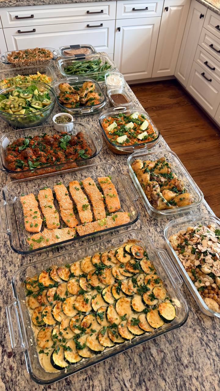 A wide spread of various glass containers filled with miso glazed salmon, Tuscan meatballs, chicken, vegetable gratin, and fresh salad on a kitchen counter.