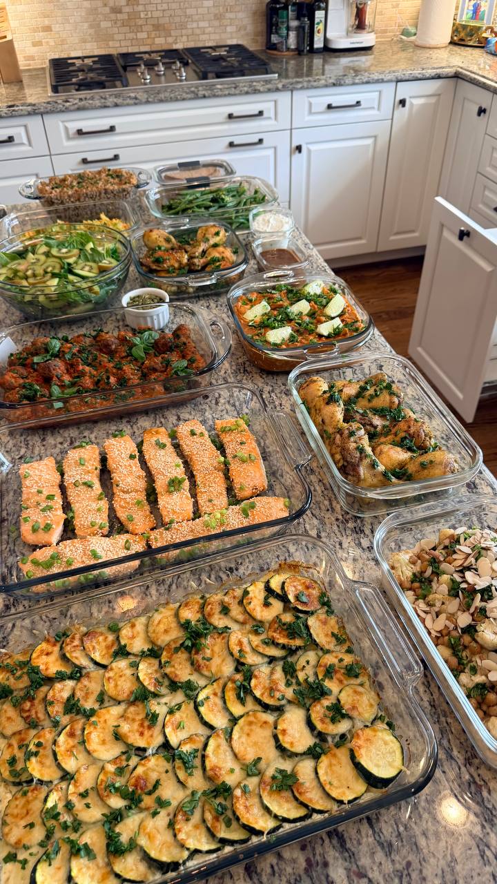 A variety of glass containers holding miso salmon, Tuscan meatballs, zucchini gratin, chicken, and salads, displayed attractively on a kitchen countertop for meal prep.
