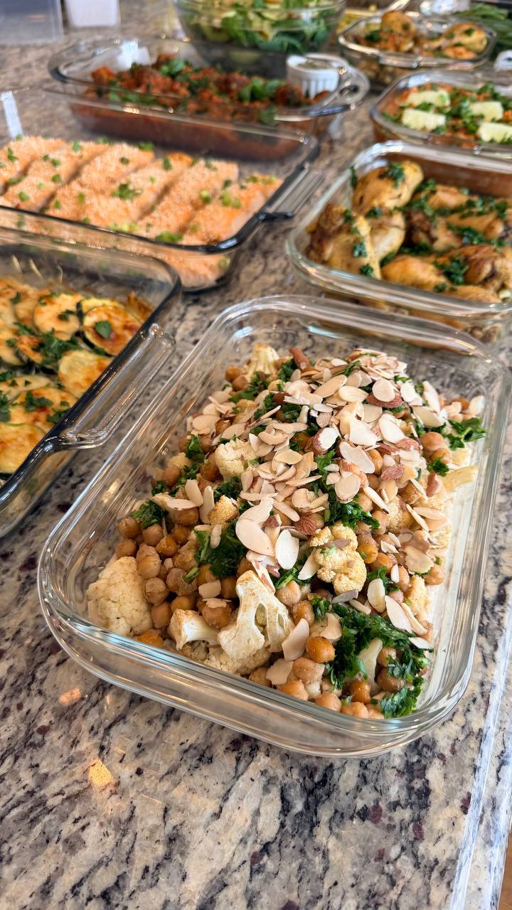 A glass container features Mediterranean spiced cauliflower, chickpeas, and fresh greens, generously topped with sliced almonds and aromatic herbs for a beautiful presentation.