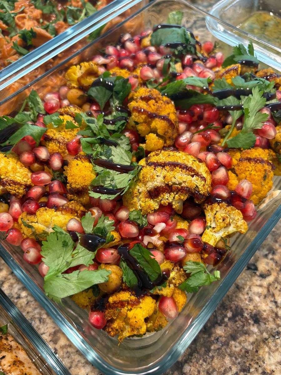 A glass container features golden roasted Mediterranean spiced cauliflower florets mixed with chickpeas, vibrant pomegranate seeds, fresh cilantro garnish, and a drizzle of balsamic glaze.