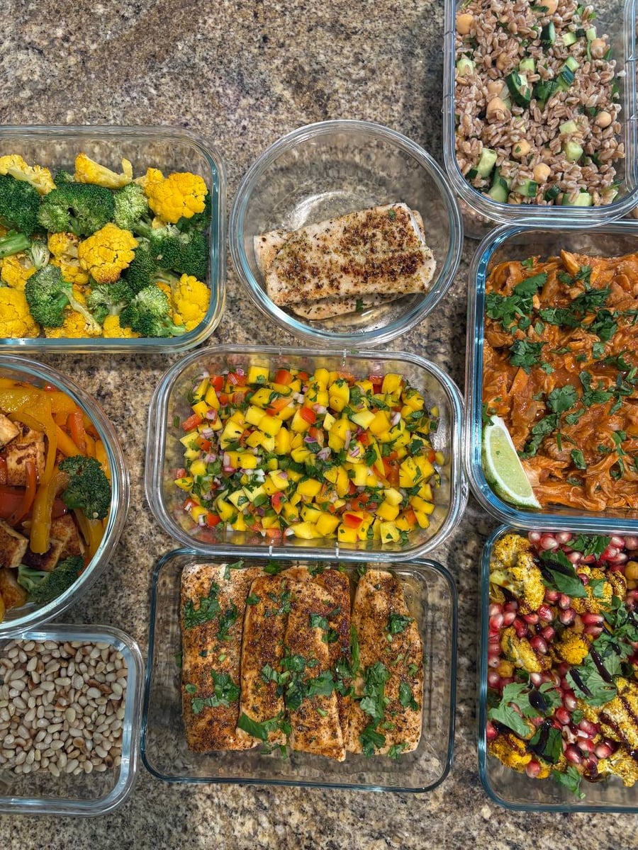 Glass meal prep containers featuring blackened mahi-mahi, mango salsa, saucy shredded chicken, herbed farro, roasted cauliflower with pomegranate, and a paneer vegetable stir-fry.