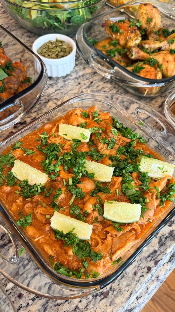 A glass container holds saucy shredded chicken thighs garnished with fresh cilantro and lime wedges, surrounded by containers of farro and cauliflower salad.