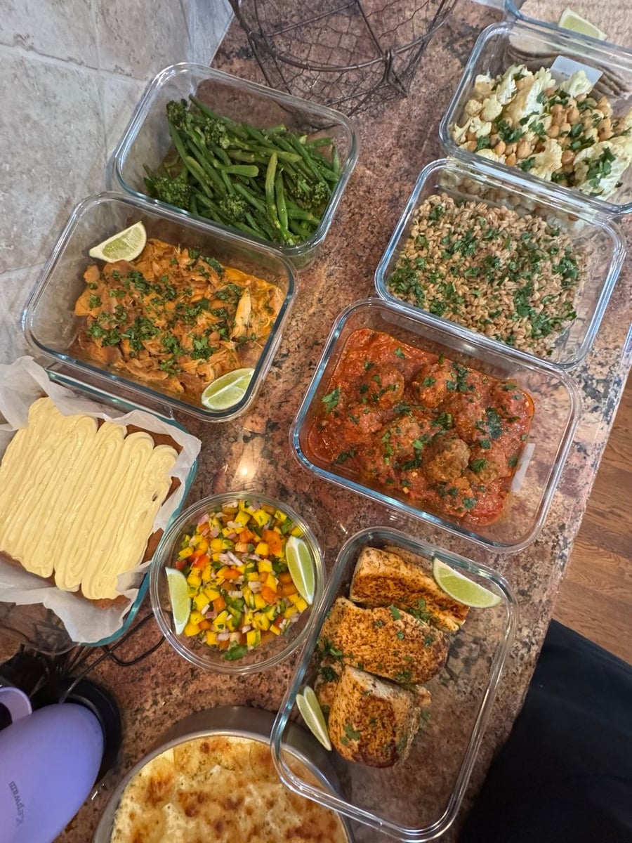 An overhead view of glass meal prep containers featuring blackened mahi mahi, Tuscan beef meatballs, shredded chicken, roasted vegetables, farro, and frosted carrot cake.