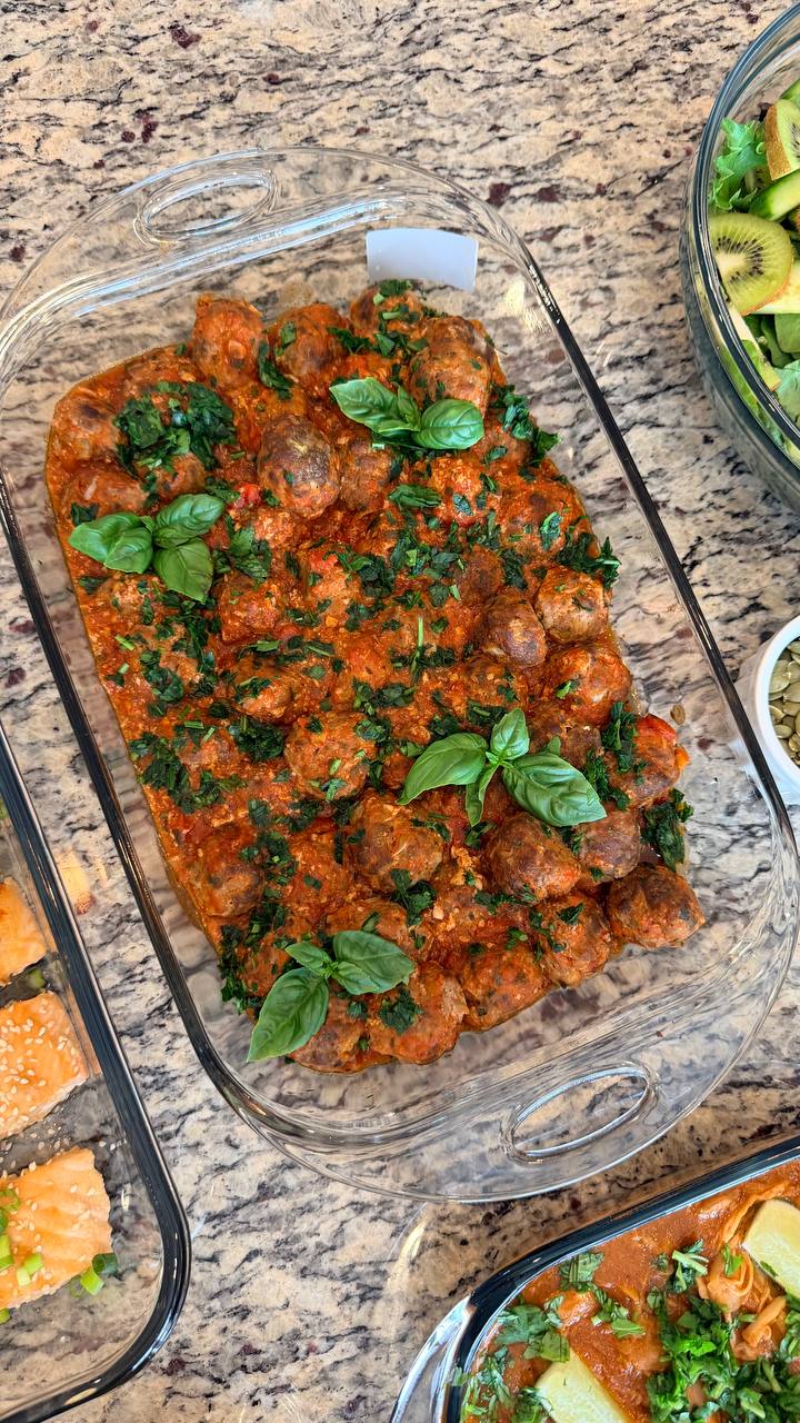 A glass baking dish filled with Tuscan-style beef meatballs poached in a vibrant tomato basil sauce, garnished with fresh whole basil leaves and chopped herbs.