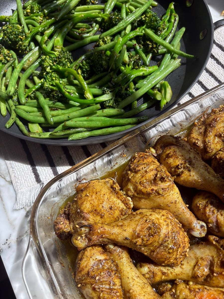 A glass baking dish with zaatar spiced chicken drumsticks alongside a skillet filled with a bright green sauté of broccoli and fresh green beans.