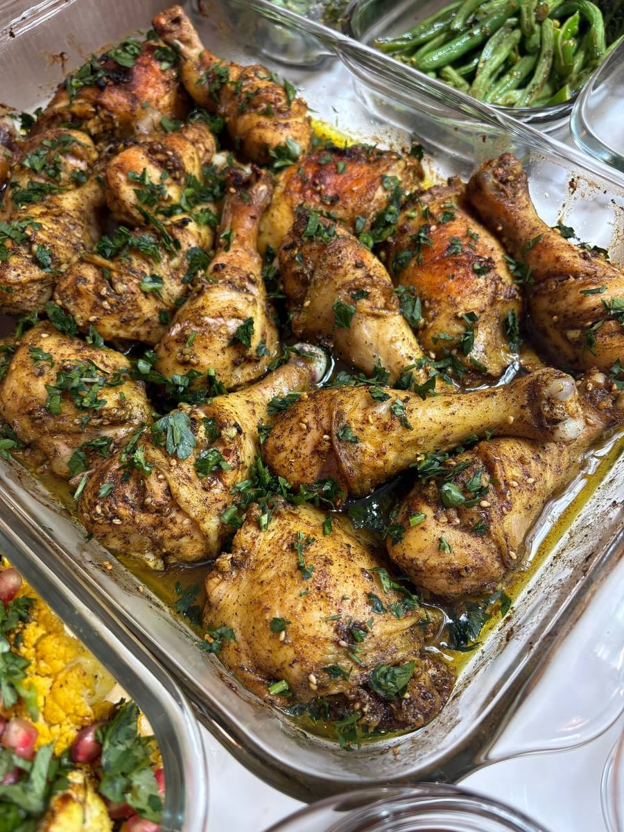 A glass baking dish filled with golden-brown zaatar spiced chicken drumsticks and thighs garnished with chopped green herbs, served alongside roasted vegetables.