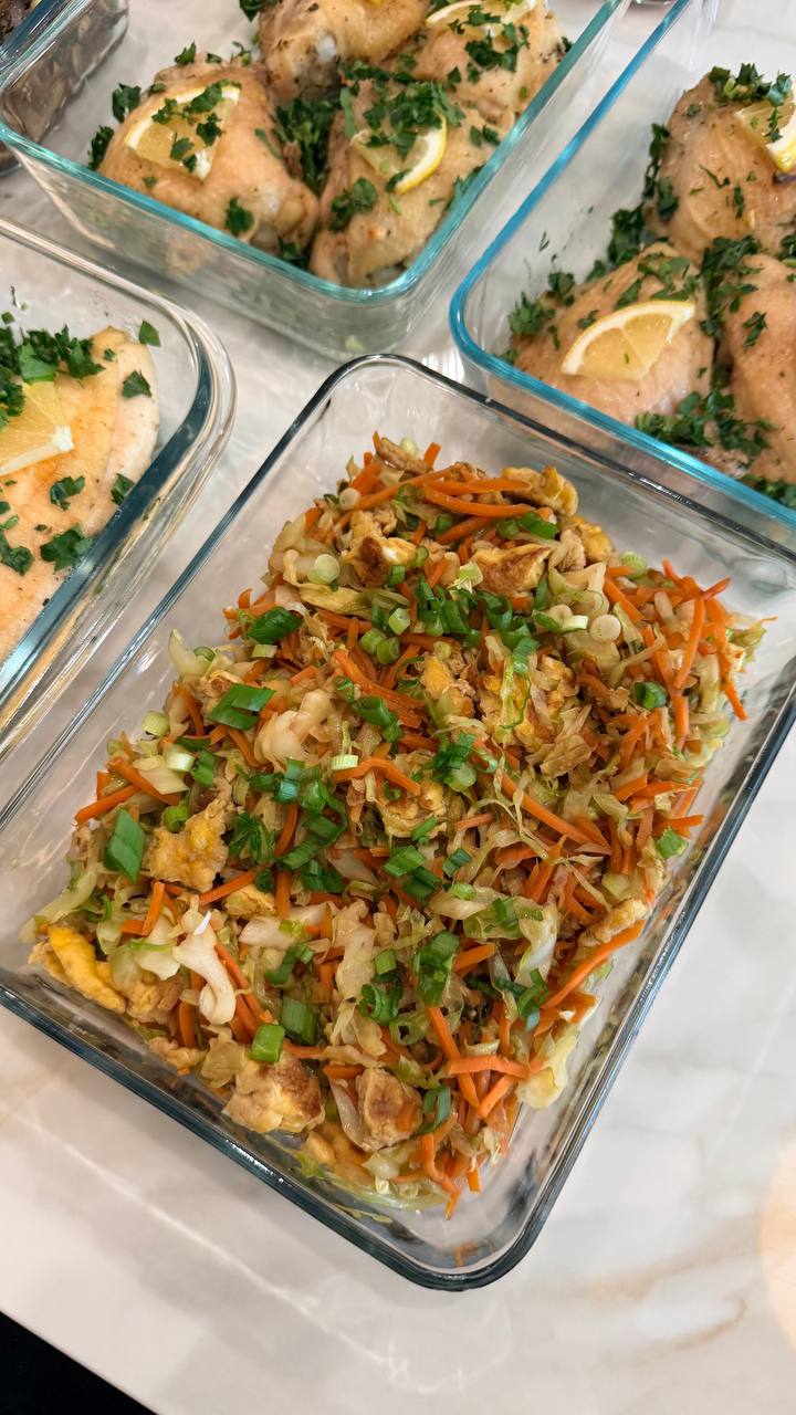 Glass meal prep containers filled with lemon baked chicken pieces and a vibrant stir-fry of cabbage, carrots, and egg, topped with fresh scallions.