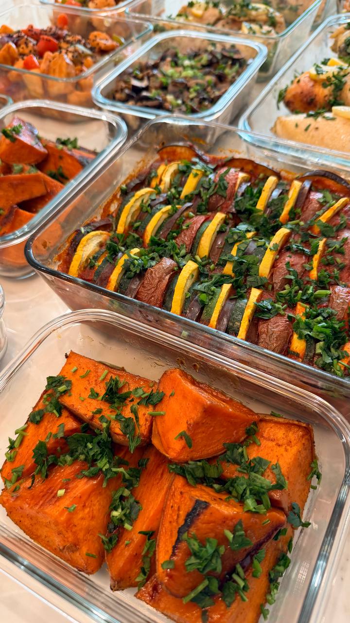 Glass meal prep containers filled with vibrant ratatouille with zucchini and squash, alongside tender baked sweet potato wedges garnished with fresh green herbs.