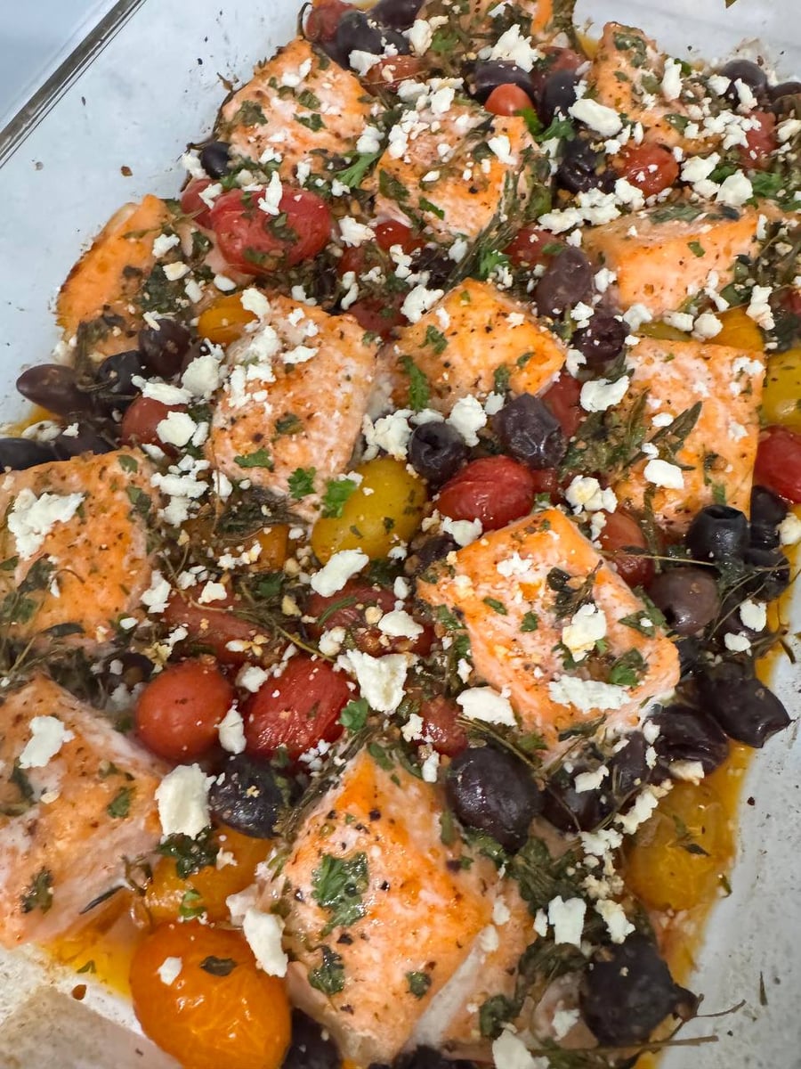 A glass baking dish filled with tender baked salmon bites, roasted cherry tomatoes, savory black olives, fresh herbs, and a generous crumbly feta cheese topping.